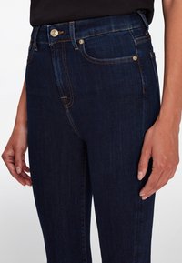 Jeans de mezclilla de un azul oscuro con corte ajustado, que cuentan con un cierre de botón, bolsillos frontales y costuras en contraste de color naranja.