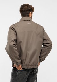 Hnedá voľná bunda s golierom, elastickými manžetami a pásom. Materiál má hladkú textúru a na zadnej strane sa nachádza diskrétne logo.