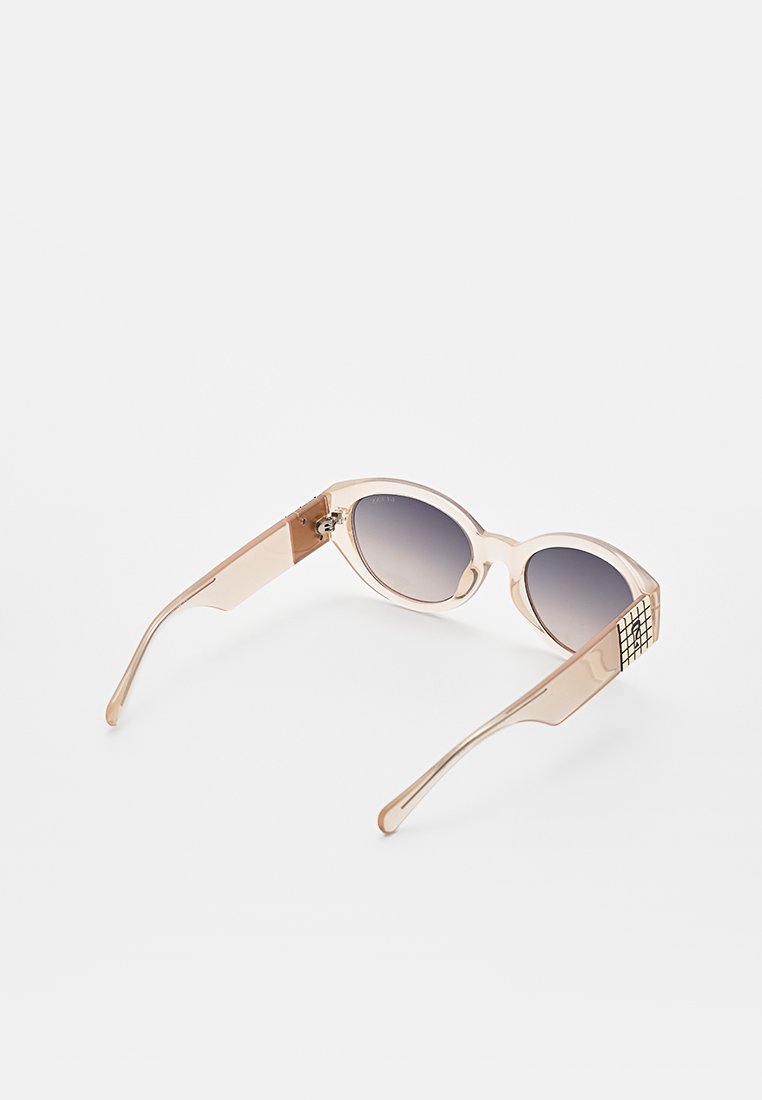 Beige durchsichtige ovale Sonnenbrille mit grauen Verlaufsgläsern und einem schwarzen Gittermuster an den Tempelenden, die auf einer weißen Oberfläche liegen.