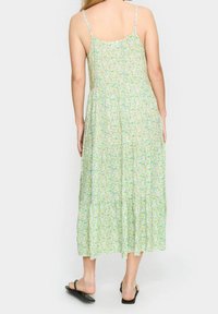 Kwiatowa maxi sukienka z cienkimi regulowanymi ramiączkami, jasna zielona ściana, a na niej wzory w kolorze różowym, żółtym i niebieskim; warstwowy, zwiewny fason.