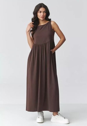 Kobieta o długich ciemnych włosach w brązowej, bezrękawnikowej sukience maxi i białych sneakersach na platformie, z jedną ręką w kieszeni, na jednolitym tle.