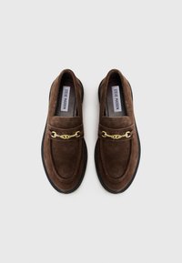 Brown suede loafers with a gold chain accent, rounded toe, and black rubber sole; features a smooth texture and stitched detailing.