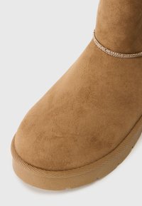Close-up of a tan suede boot toe with stitched trim and textured rubber sole on a white background.