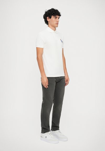 Camisa polo branca com colarinho, combinada com jeans cinza escuro e ténis brancos. A polo apresenta um pequeno design bordado no peito.