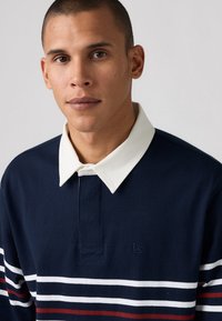 Polo de manga larga en color navy con cuello blanco; presenta rayas horizontales en blanco, rojo y navy; logo bordado en el lado izquierdo.