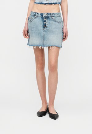 Woman wearing a light blue button-front denim mini skirt with frayed hem, paired with black pointed flats and a cropped denim top.