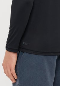 Czarna koszula z długim rękawem wykonana z gładkiego materiału. Posiada metkę UPF 50+ przy dolnym brzegu. Projekt zawiera płaskie szwy oraz dopasowany krój.