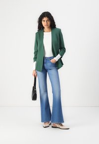 Zelený blazer se strukturovaným designem, bílý top s dlouhým rukávem, rozšířené modré džíny a černá kabelka. Boty jsou béžové baleríny se pásem.