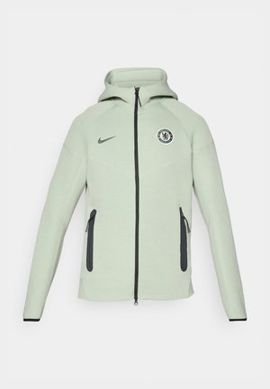Hele roheline kapuutsiga jakk, millel on lukuga sulgemine, ümmargune Chelsea FC logo, mustad aksessuaarid ja kaks eesmist lukuga taskut.