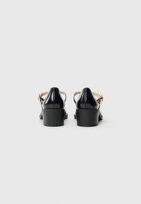 Black patent leather women's block heel shoes with ankle straps and gold buckle, viewed from the back on a white background.