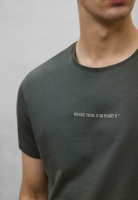 Mørkegrøn bomulds T-shirt med rund halsudskæring og korte ærmer. Har hvid tekst: "FORDI DER IKKE ER EN PLANET B" trykt på forsiden.
