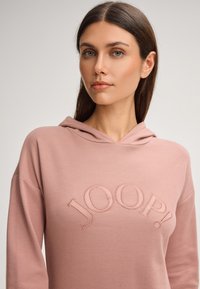 Jasnoróżowa bluza z kapturem wykonana z miękkiego materiału; posiada duży, haftowany napis "JOOP!" w kształcie łuku. Miękka tekstura, luźny krój i sznurek do regulacji kaptura.