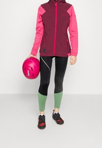Chaqueta con capucha en color marrón y rosa con cremallera, leggings negros con acentos verdes, y zapatillas de ciclismo negras sosteniendo un casco rosa.
