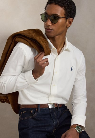 Мъж, носещ слънчеви очила, бяла риза с копчета, сини дънки и кафяв колан, държащ кафяво яке на рамото си пред обикновен фон.