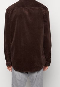 Hnědá manšestrová košile se zadním dílem, manžetami na knoflíky, zakřiveným lemem a jemnou texturou. Má volný střih a je kombinována se světle šedými kalhotami.