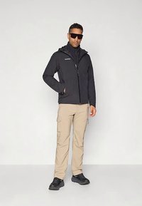 Mężczyzna w czarnej kurtce z kapturem, beżowych spodniach typu cargo, czarnych butach i ciemnych okularach przeciwsłonecznych, stojący na tle jasnego, jednolitego tła.