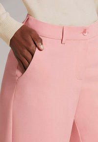 Lichtroze broek van gladde stof, met een zijzak, opgerolde tailleband en een enkele knoopsluiting.