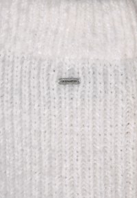 Weißer Strickbeanie aus flauschigem Garn mit geripptem Muster. Ein kleines silbernes Etikett mit "DREIMASTER" ist an der Seite angenäht.