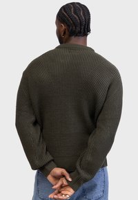 Suéter tejido en verde oliva con un patrón texturizado, hombros caídos y puños acanalados. La parte trasera presenta un diseño de costura único.
