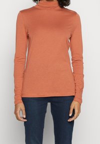 Långärmad polotröja i roströd färg, gjord av mjukt material. Har en figursydd design och en rak fåll, i kombination med mörkblå jeans.