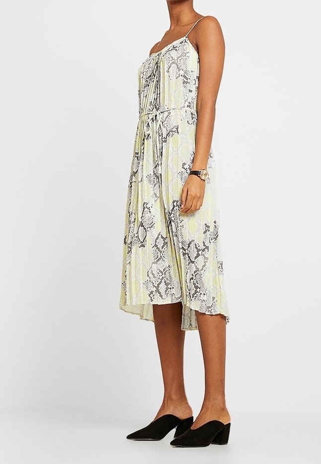 Femme portant une robe midi sans manches à imprimé serpent avec fines bretelles, des mules noires et une montre au poignet, debout devant un fond uni.