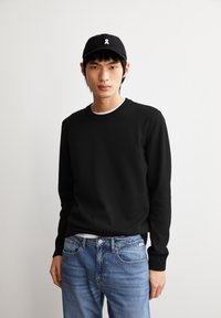 Schwarzes Langarm-Sweatshirt mit geripptem Ausschnitt, über einem weißen Shirt getragen, kombiniert mit hellblauen Denim-Jeans. Schwarze Kappe mit weißem Logo.