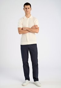 Kortärmad stickad beige pikétröja med en strukturerad ribbad mönstring. Tillsammans med marinblå chinos och vita sneakers, som står framför en ljus bakgrund.