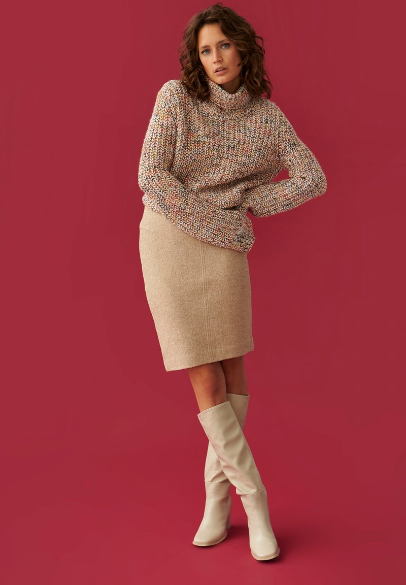 Suéter de punto en multicolor con un cuello alto, combinado con una falda lápiz beige y botas de crema hasta la rodilla, todo sobre un fondo rojo.