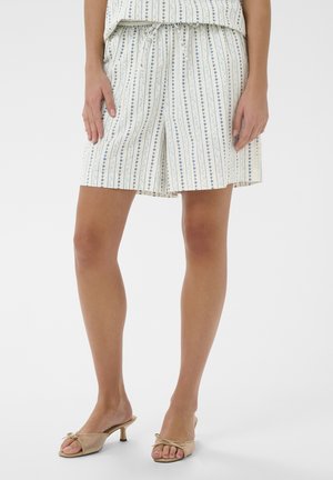 Kvinde iført hvide shorts med lodrette blå blomsterstriber, en matchende top og beige åbne hælede sandaler med sløjfer.
