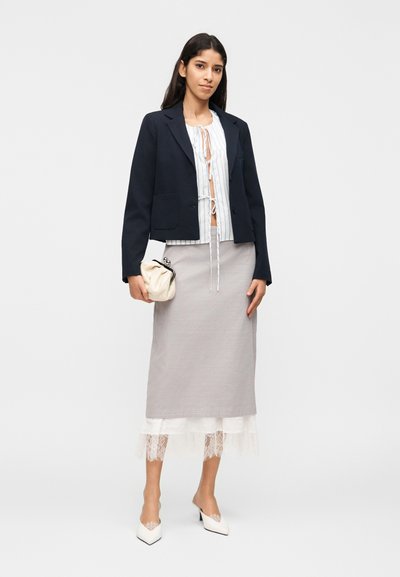 Γυναίκα με ναυτικό μπλέ σακάκι, ριγέ πουκάμισο με δέσιμο μπροστά, γκρι φούστα με λευκή δαντέλα στο τελείωμα, λευκά μυτερά τακούνια, κρατώντας κρεμ τσάντα φάκελο.