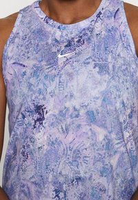 Gros plan d'une personne portant un haut sans manches Nike de couleur violette à motif abstrait, avec un logo Swoosh blanc sur la poitrine.