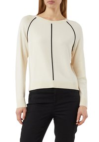 Frau trägt einen cremefarbenen Pullover mit langen Ärmeln und schwarzen, vertikalen sowie geschwungenen Abnähern, kombiniert mit schwarzen, enganliegenden Hosen.