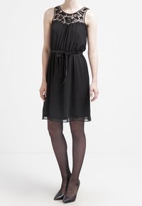 Robe noire sans manches avec un détail en dentelle au niveau du décolleté, taille froncée et jupe fluide. Portée avec des collants noirs transparents et des talons aiguilles.