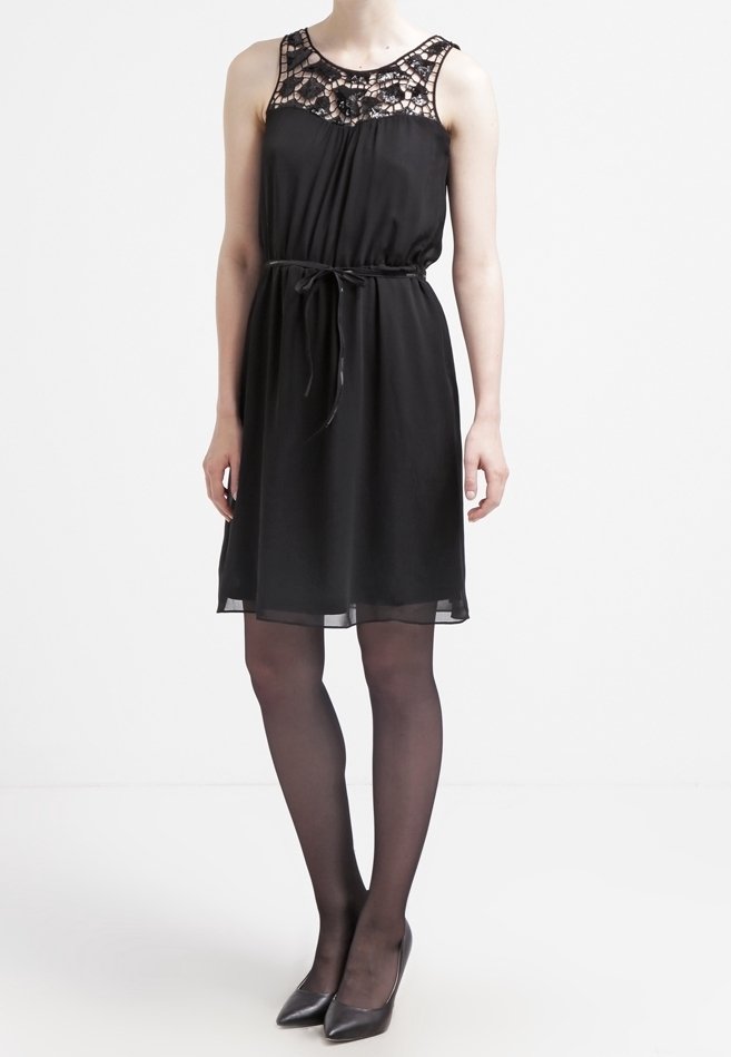 Robe noire sans manches avec un détail en dentelle au niveau du décolleté, taille froncée et jupe fluide. Portée avec des collants noirs transparents et des talons aiguilles.