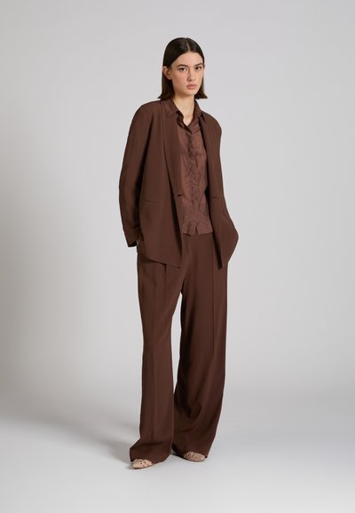 Frau steht in locker sitzendem braunem Anzug mit passendem Hemd und beigen Schuhen vor einem schlichten grauen Hintergrund.