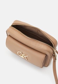 Světle hnědá kožená crossbody taška se zlatým zipem a detaily loga CK, částečně otevřená, ukazující interiér s potištěným podšívkou.