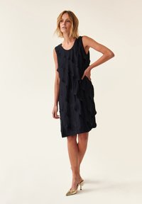 Robe noire sans manches avec des volants en cascade, longueur genoux et tissu léger ; portée avec des chaussures à talons hauts de couleur dorée.