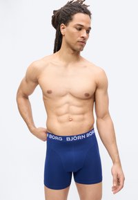 Homme torse nu avec des dreadlocks portant un boxer bleu Björn Borg, debout avec une main sur la hanche, regardant vers le bas sur le côté.