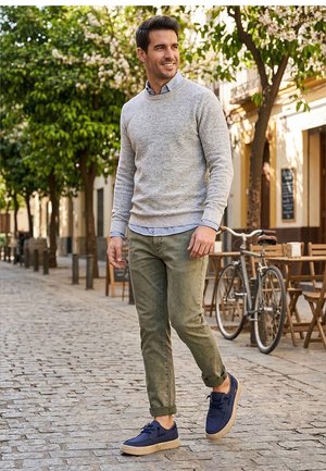 Hombre caminando por una calle adoquinada con un suéter gris, pantalones verdes y zapatos azul marino, con una cafetería al aire libre y una bicicleta cerca.