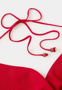 Tissu rouge avec un cordon fin noué comportant deux perles en bois aux extrémités, posé sur une surface blanche unie.