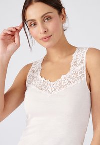Top sin mangas de color beige claro con un escote de encaje en forma de ola. Presenta una tela suave y lisa y un diseño ajustado.