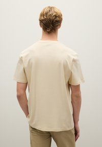 T-shirt beige a maniche corte con collo rotondo, realizzato in tessuto morbido. Presenta una vestibilità rilassata e una texture liscia, design minimalista.