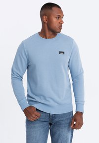 Sudadera azul clara con cuello redondo, puños y dobladillo acanalados. Presenta un parche de logo rectangular negro en el pecho. Combinada con jeans azules.