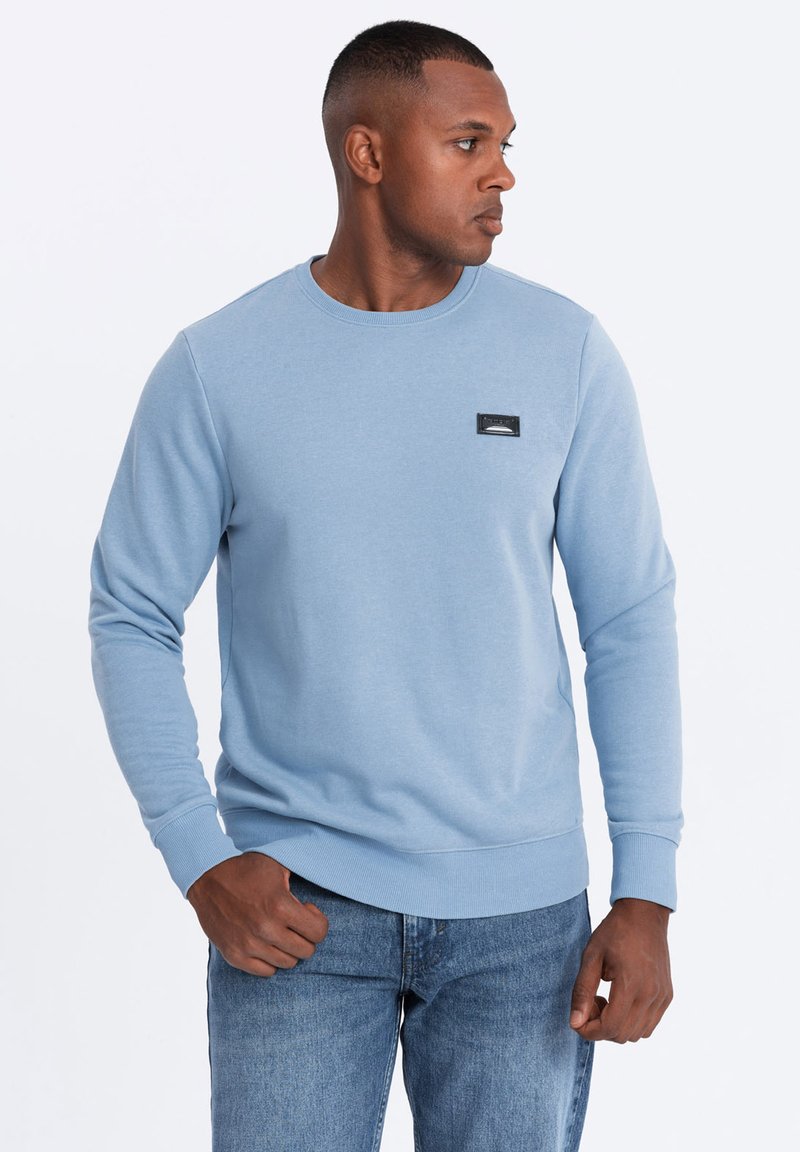 Sudadera azul clara con cuello redondo, puños y dobladillo acanalados. Presenta un parche de logo rectangular negro en el pecho. Combinada con jeans azules.