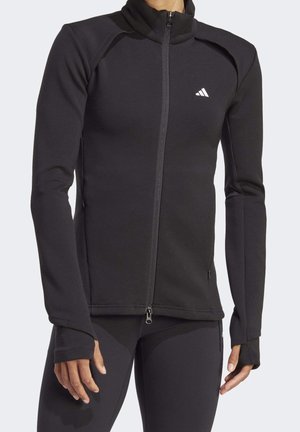 Osoba nosi dopasowaną czarną kurtkę na zamek z otworami na kciuki i małym białym logo Adidas na piersi.