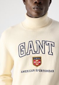 Cremfarbener Rollkragenpullover mit navyfarbenem "GANT"-Text und einem roten und gelben Logopatch. Gestrickte Textur mit geripptem Kragen und Bündchen.