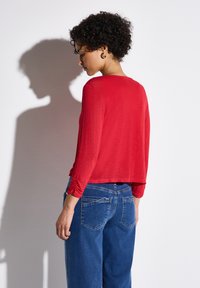 Röd långärmad topp med rund halsringning och rynkade ärmdetaljer, tillsammans med löst sittande blå denimjeans. Skugga kastad på väggen.