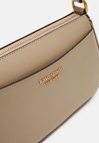 Beige crossbody bag made of textured material, featuring a gold zipper, gold logo accent, and a slim design with a flat pocket.