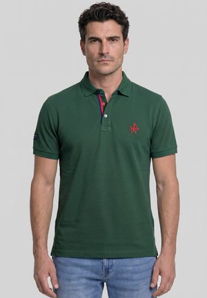Hombre con una camiseta tipo polo de manga corta verde oscuro con detalles rojos y azules en la tapeta interior y vaqueros azul claro sobre un fondo liso.
