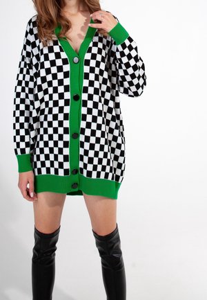 Femme portant un cardigan à carreaux noir et blanc avec un bord vert et des boutons, associé à des bottes noires montantes jusqu'au genou sur fond blanc.